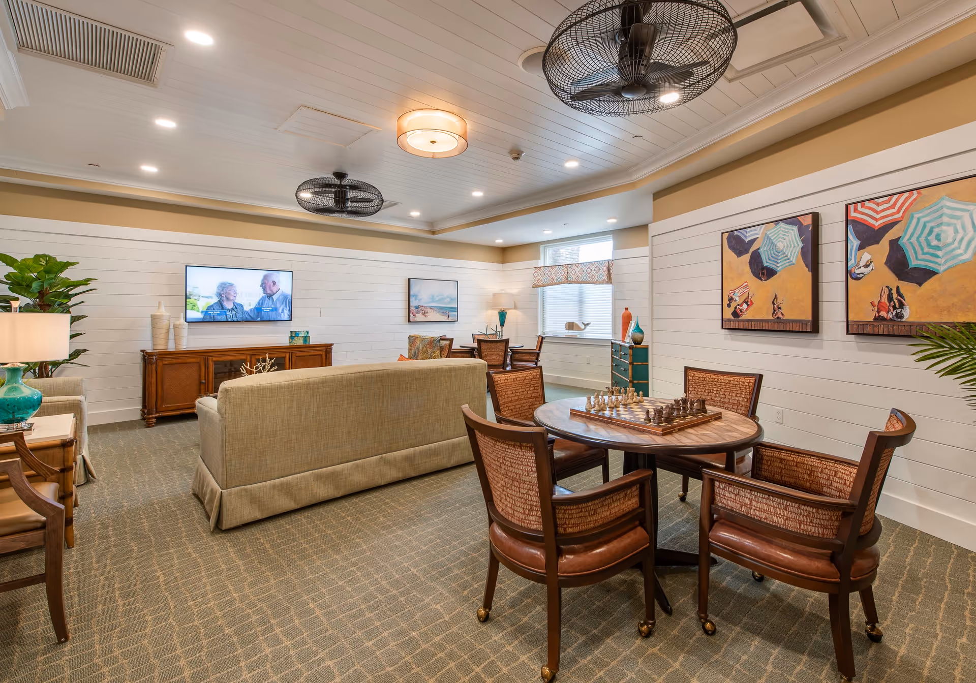 A cozy living room area in a senior living facility with beige sofas facing a wall-mounted TV, a round wooden table with a chess set and four chairs, decorative paintings on the walls, and ceiling fans. The room has carpeted flooring and soft lighting.