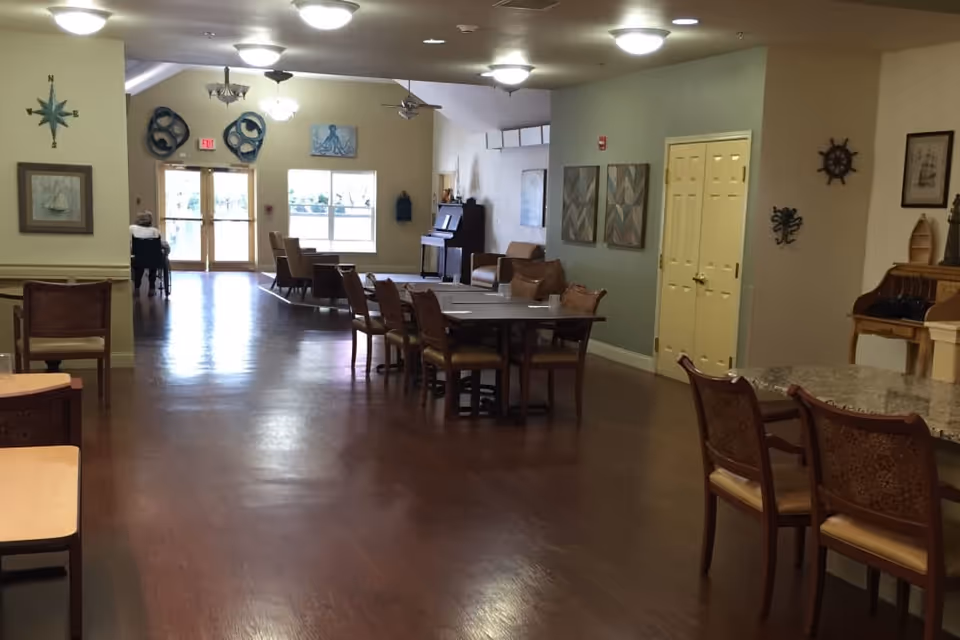 Interior view of a senior living facility common area with wooden floors, multiple tables and chairs, wall decorations including nautical-themed items, a piano, and a person in a wheelchair near the exit doors.