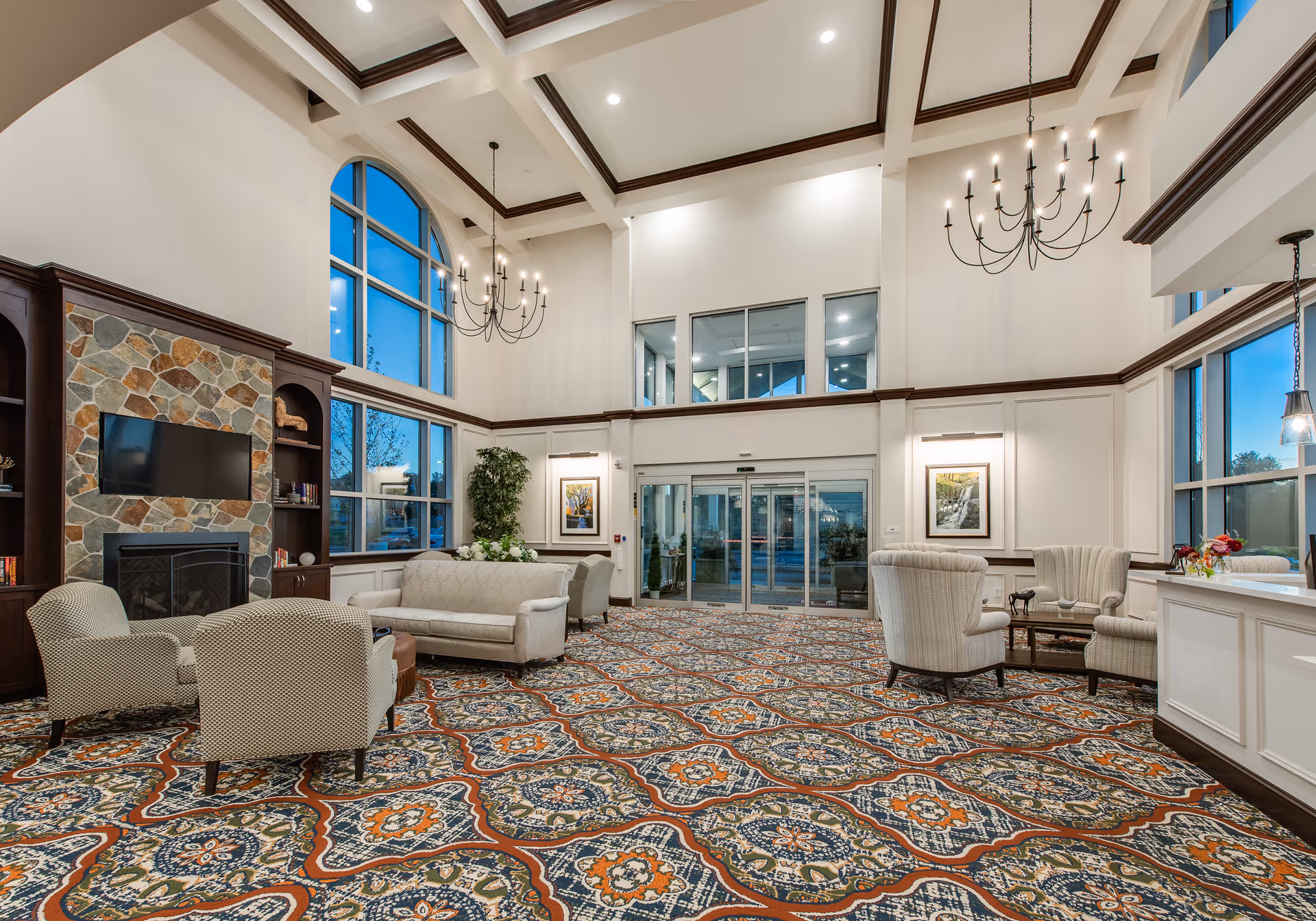 Spacious and elegant senior living facility lobby with high ceilings, large windows, and chandeliers. The room features a patterned carpet, a stone fireplace with a mounted TV, comfortable seating including sofas and armchairs, and a reception desk on the right side.