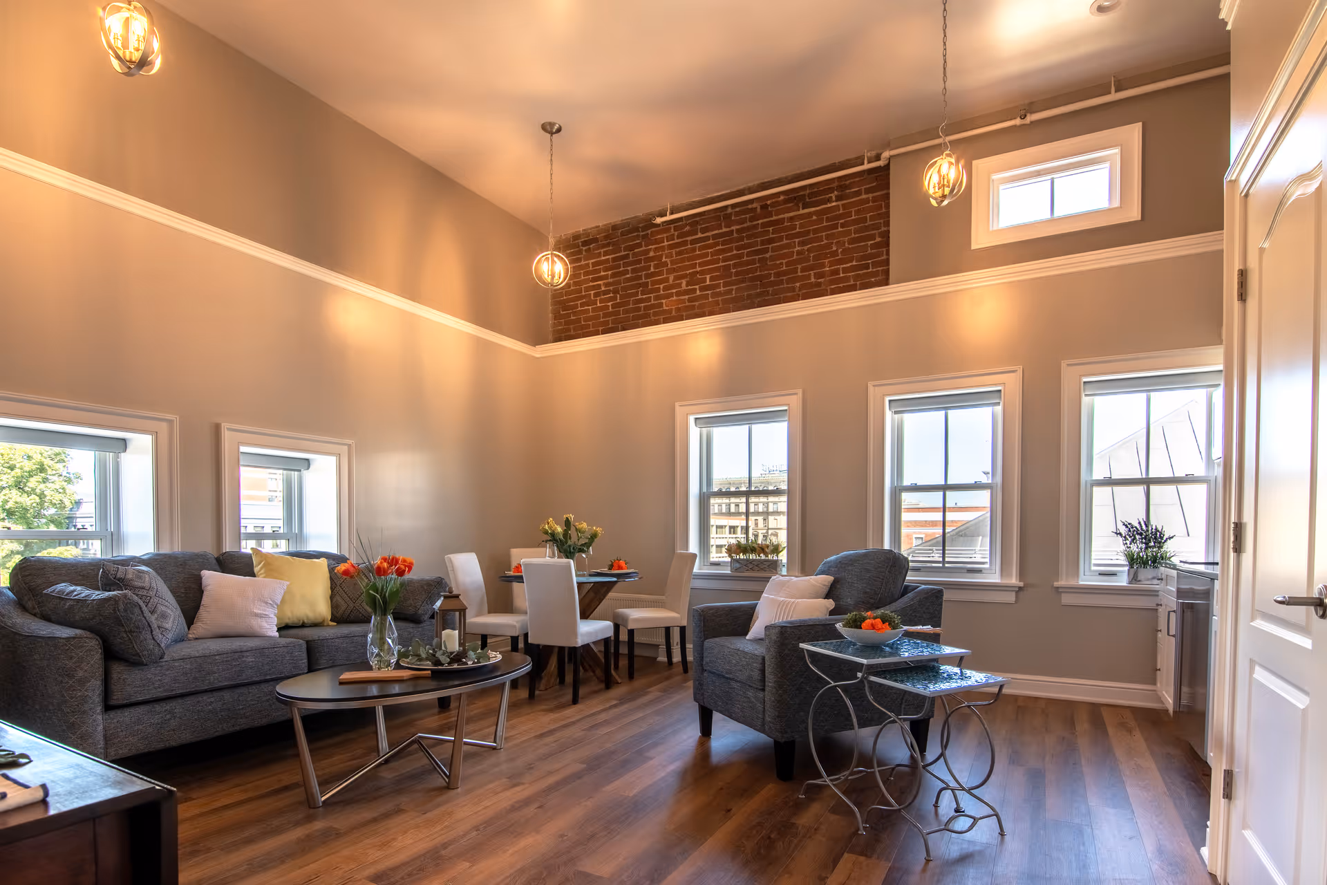 A bright and spacious living room with high ceilings and multiple windows letting in natural light. The room features a gray sofa with colorful cushions, a matching armchair, a round coffee table with a vase of flowers, and a small dining table with four white chairs. The walls are painted light gray with a brick accent near the ceiling, and the floor is wooden.