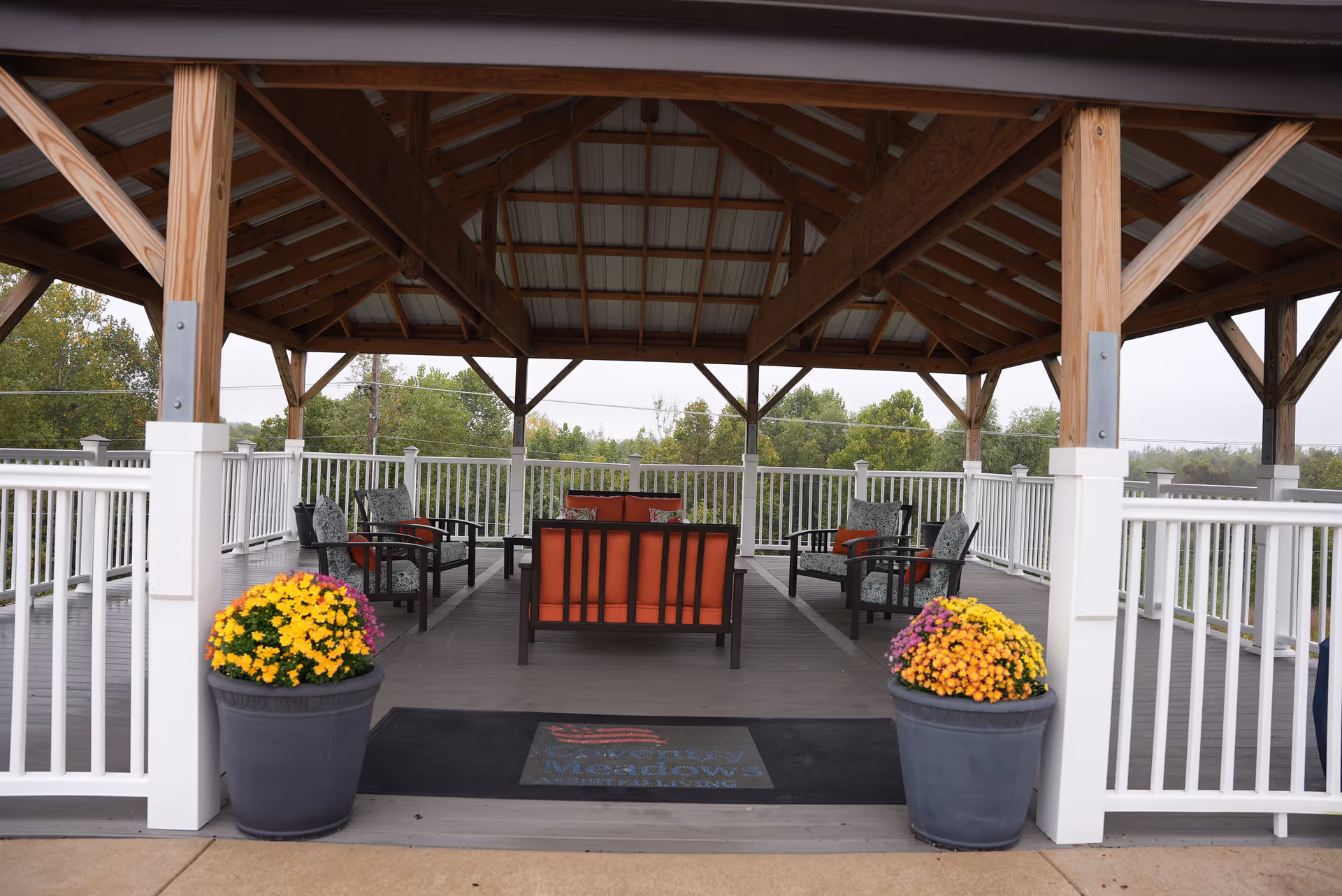 Covered outdoor seating area with wooden beams and a metal roof, featuring cushioned chairs and a loveseat arranged around a central space. Two large flower pots with colorful flowers flank the entrance, and white railings surround the deck. Trees and greenery are visible in the background.