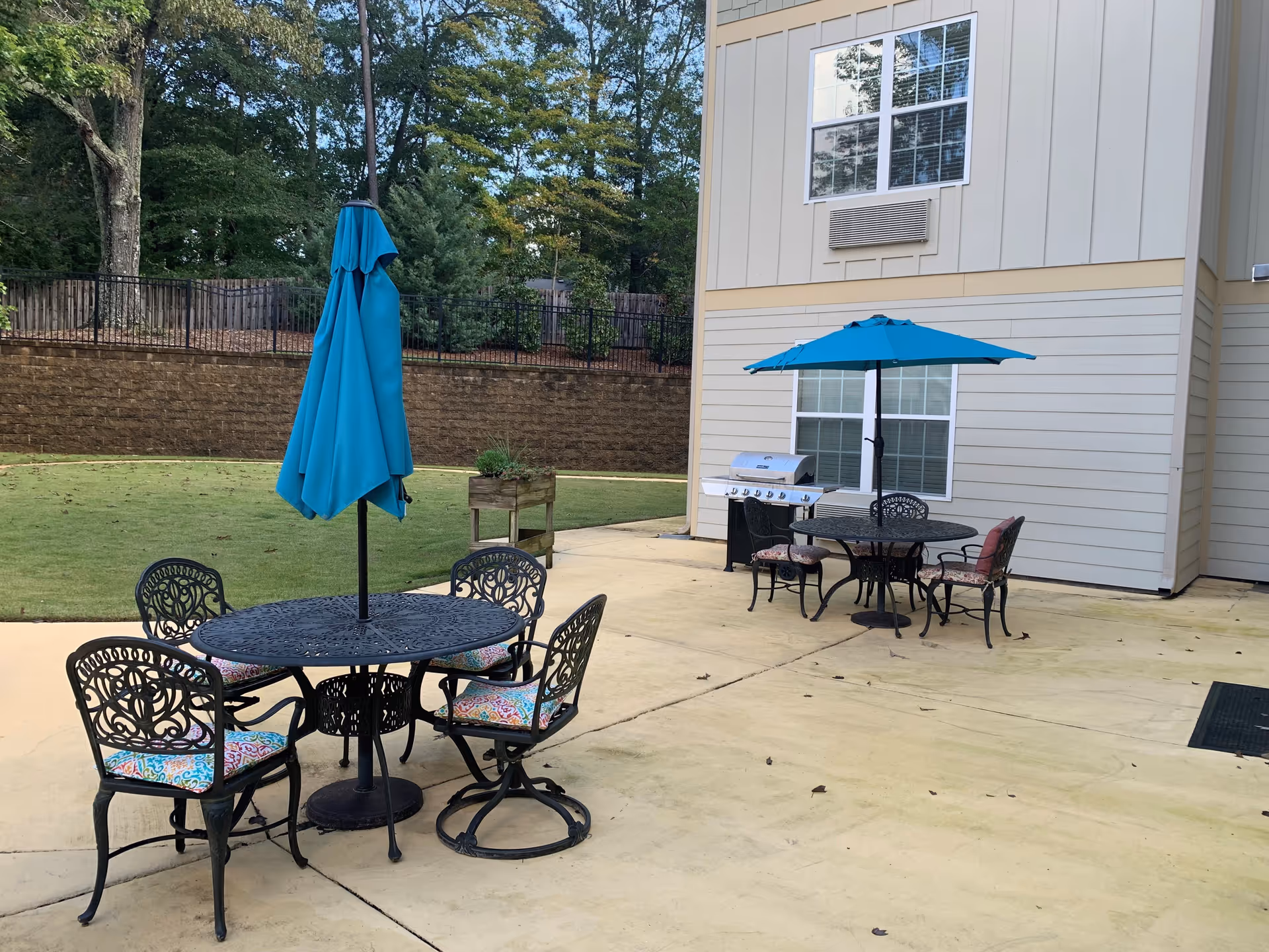 Outdoor patio area with two black metal tables, each with a blue umbrella. Four chairs with colorful cushions surround the table in the foreground, and three chairs with cushions surround the table in the background. A barbecue grill is positioned near the building wall. The area is paved and adjacent to a grassy lawn with trees and a retaining wall in the background.