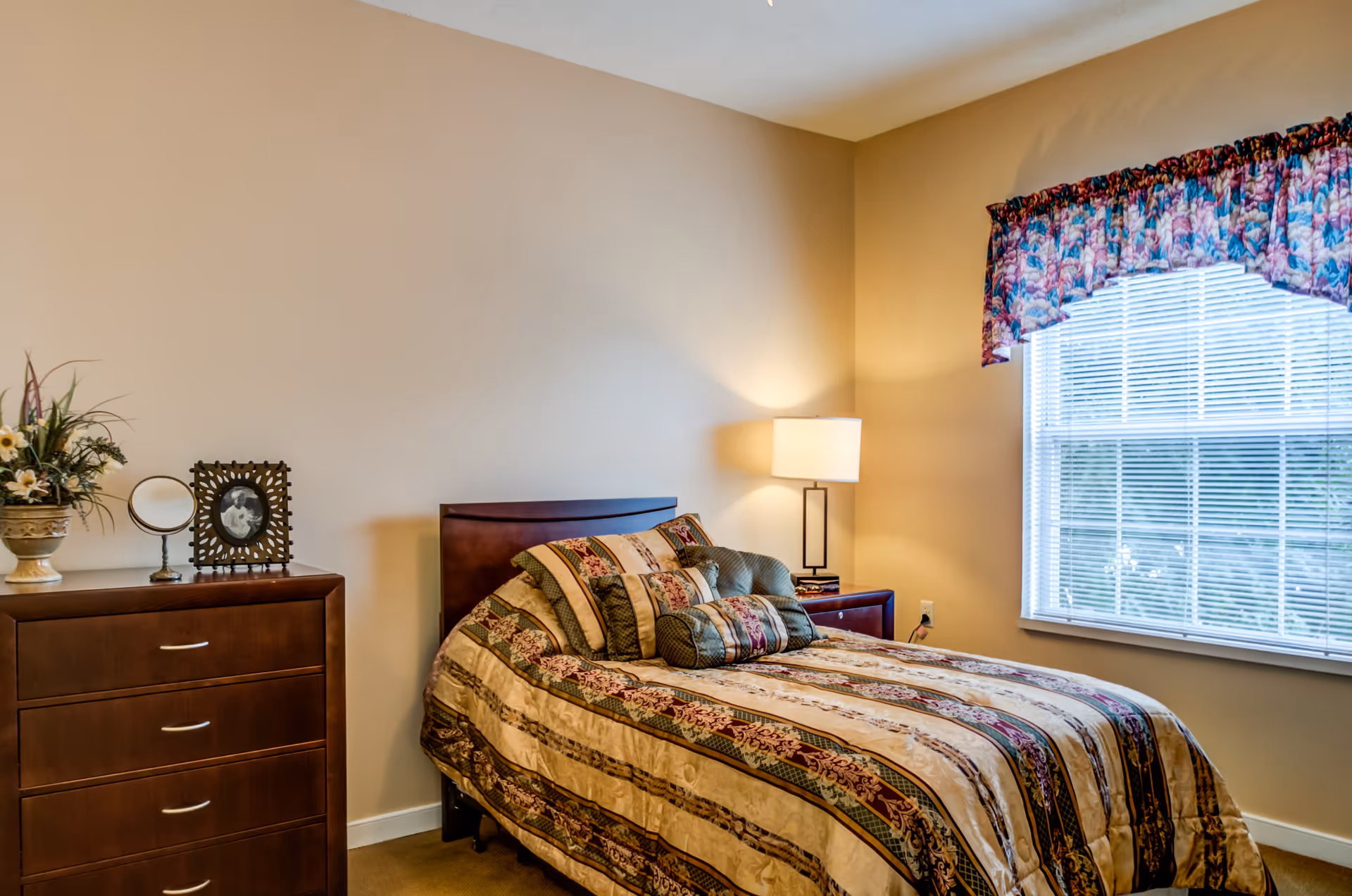 A cozy bedroom with a single bed covered in a patterned quilt and multiple pillows. Next to the bed is a wooden nightstand with a lamp and books. A wooden dresser with a flower vase, a small mirror, and a framed photo is positioned against the wall. A window with blinds and a floral valance allows natural light into the room.