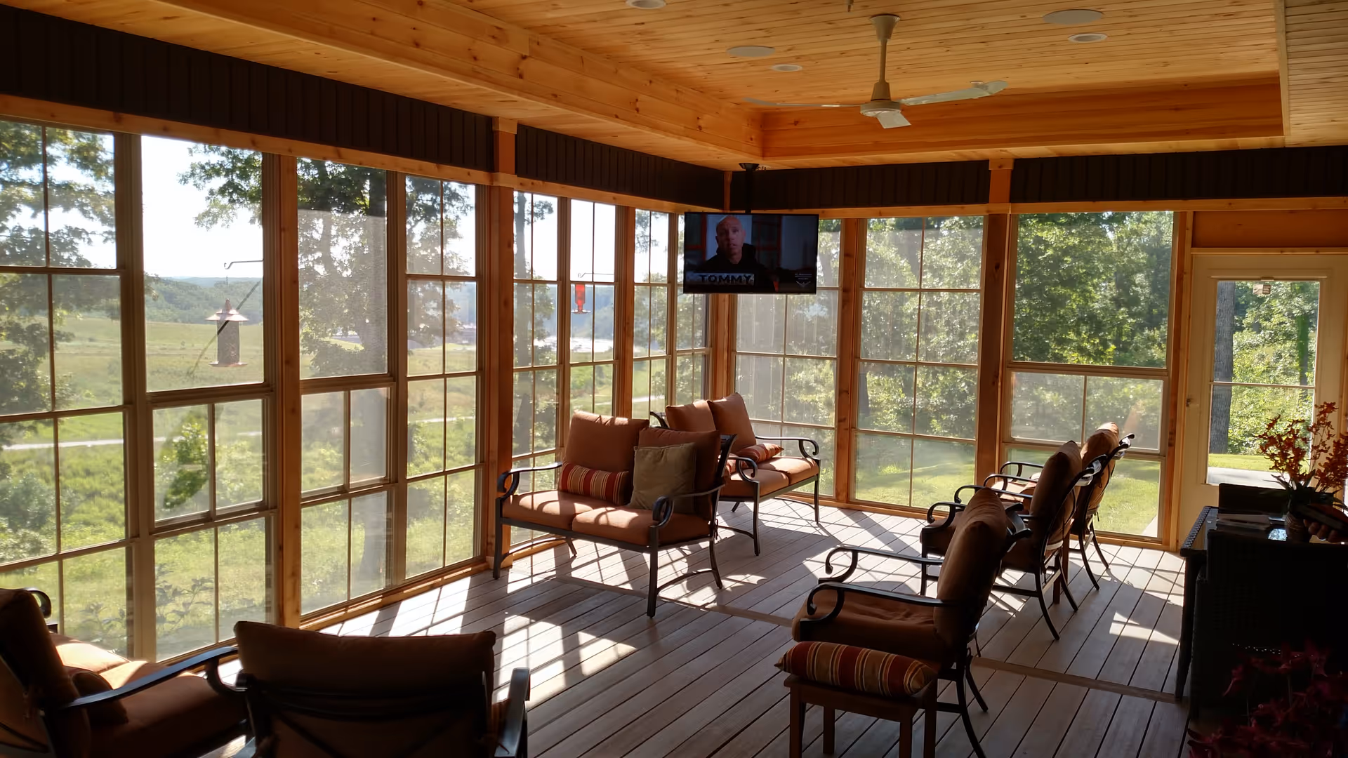 Sunlit enclosed sunroom with a wooden ceiling, floor-to-ceiling windows, cushioned armchairs and a wall-mounted TV overlooking green lawns.