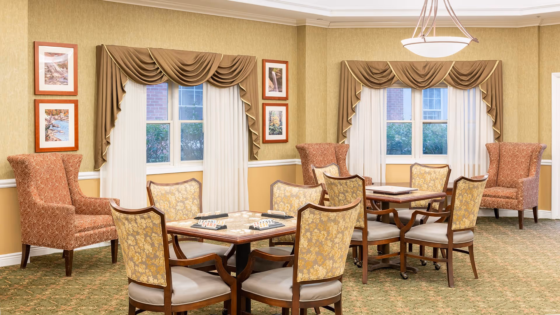 A well-lit common area with two square tables, each surrounded by four floral-patterned chairs. The tables have game pieces on them, suggesting a recreational space. The room features two large windows with brown drapes and white sheer curtains. There are two upholstered armchairs with a red and beige pattern placed near the windows. The walls are painted in a warm yellow tone with white trim, and framed artwork hangs on the walls. A ceiling light fixture hangs above the tables.