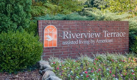 A brick sign for Riverview Terrace assisted living by Americare surrounded by green bushes and colorful flowers with trees in the background.