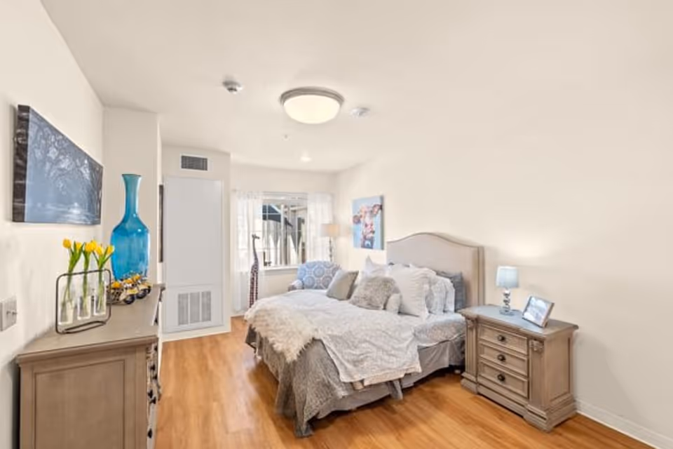 A bright and cozy bedroom with a large bed covered in white and gray bedding and multiple pillows. There is a wooden nightstand with a small lamp and a framed photo on the right side of the bed. On the left side of the room, there is a wooden dresser with a blue vase, a small flower arrangement, and a decorative item. A window with sheer white curtains allows natural light to fill the room. The floor is wooden, and the walls are painted white. A piece of artwork hangs above the bed, and a comfortable armchair with a patterned cushion is placed near the window.