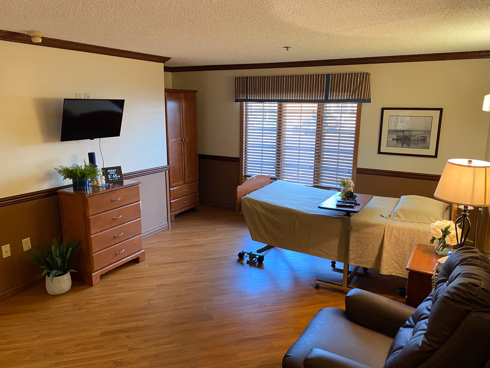 Furnished senior living bedroom with an adjustable bed and overbed table, dresser with wall-mounted TV, recliner, and large window with blinds.