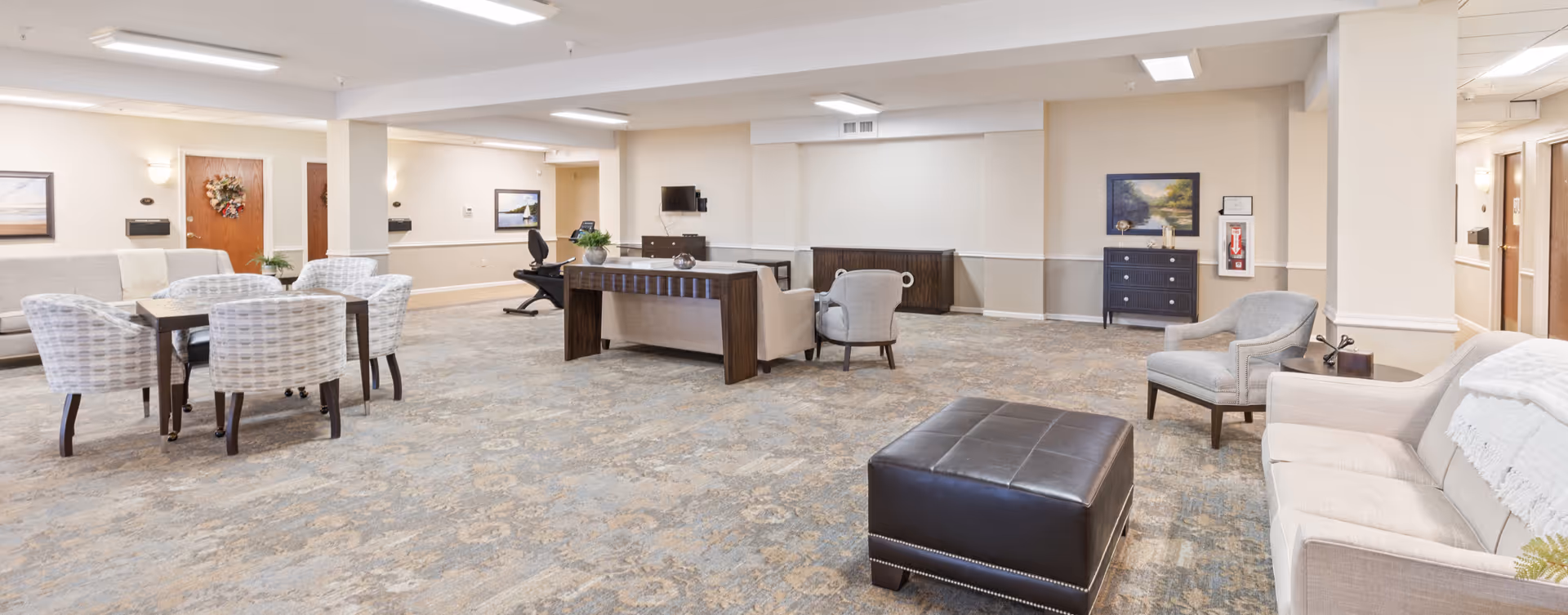 A spacious senior living facility common area with beige walls and carpeted floor. The room features multiple seating arrangements including a dining table with six cushioned chairs, several armchairs, a sofa, and a large dark ottoman. There is a desk with a chair and a wall-mounted TV in the background. The room is well-lit with ceiling lights and decorated with framed artwork and a wreath on a door.