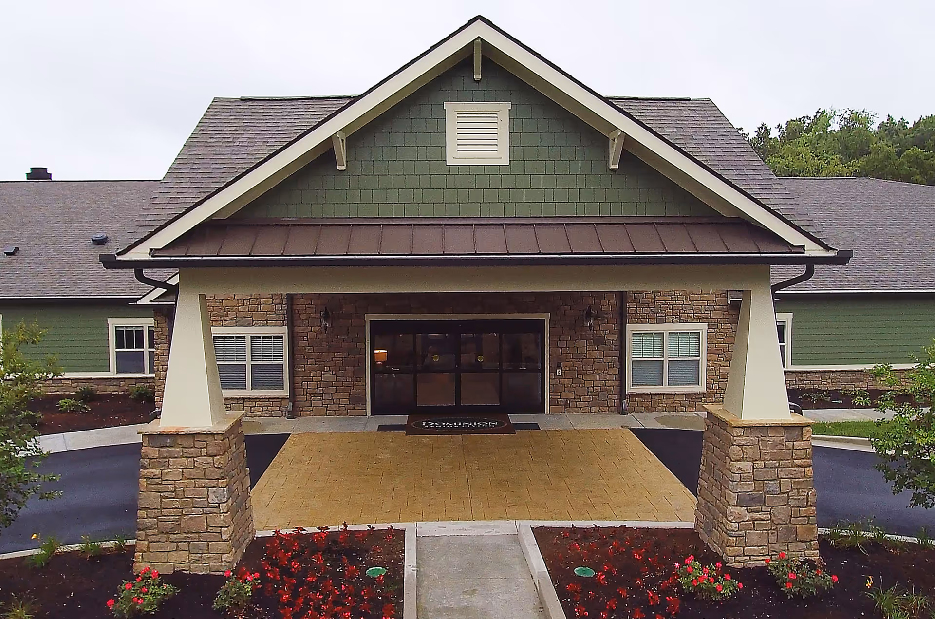 Front entrance of Dominion Senior Living of Crossville featuring a covered driveway with stone pillars, green siding, and a stone facade around the door. There are flower beds with red flowers on either side of the walkway leading to the entrance.