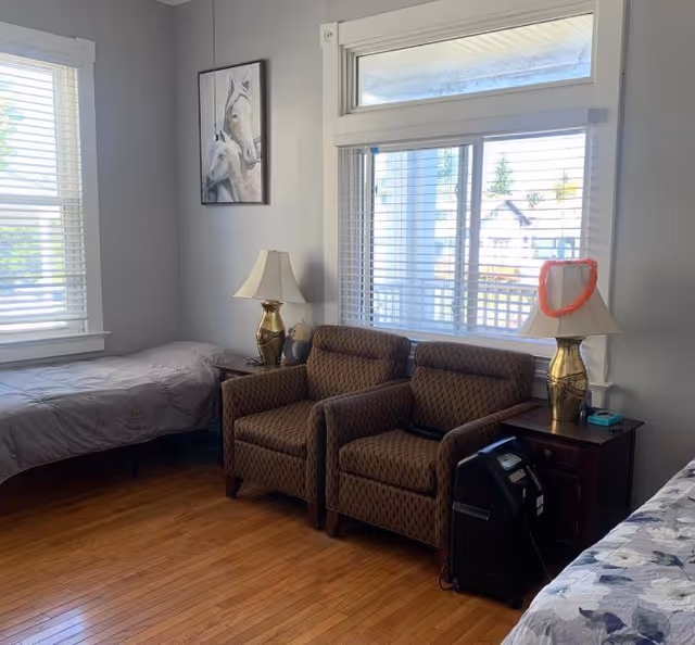 A cozy room with two single beds on opposite sides, two patterned armchairs between the beds, two side tables each with a lamp, a painting of horses on the wall, and large windows with blinds letting in natural light.