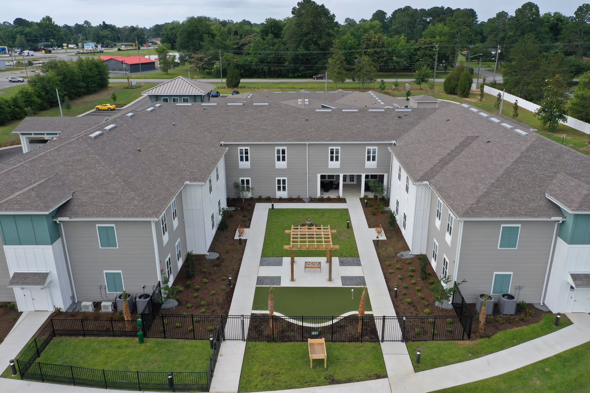 Aerial view of a senior living facility building with a U-shaped layout surrounding a landscaped courtyard featuring a small pergola, walking paths, and green lawn. The building has gray siding with white trim and multiple windows. There are trees and a parking area visible in the background.