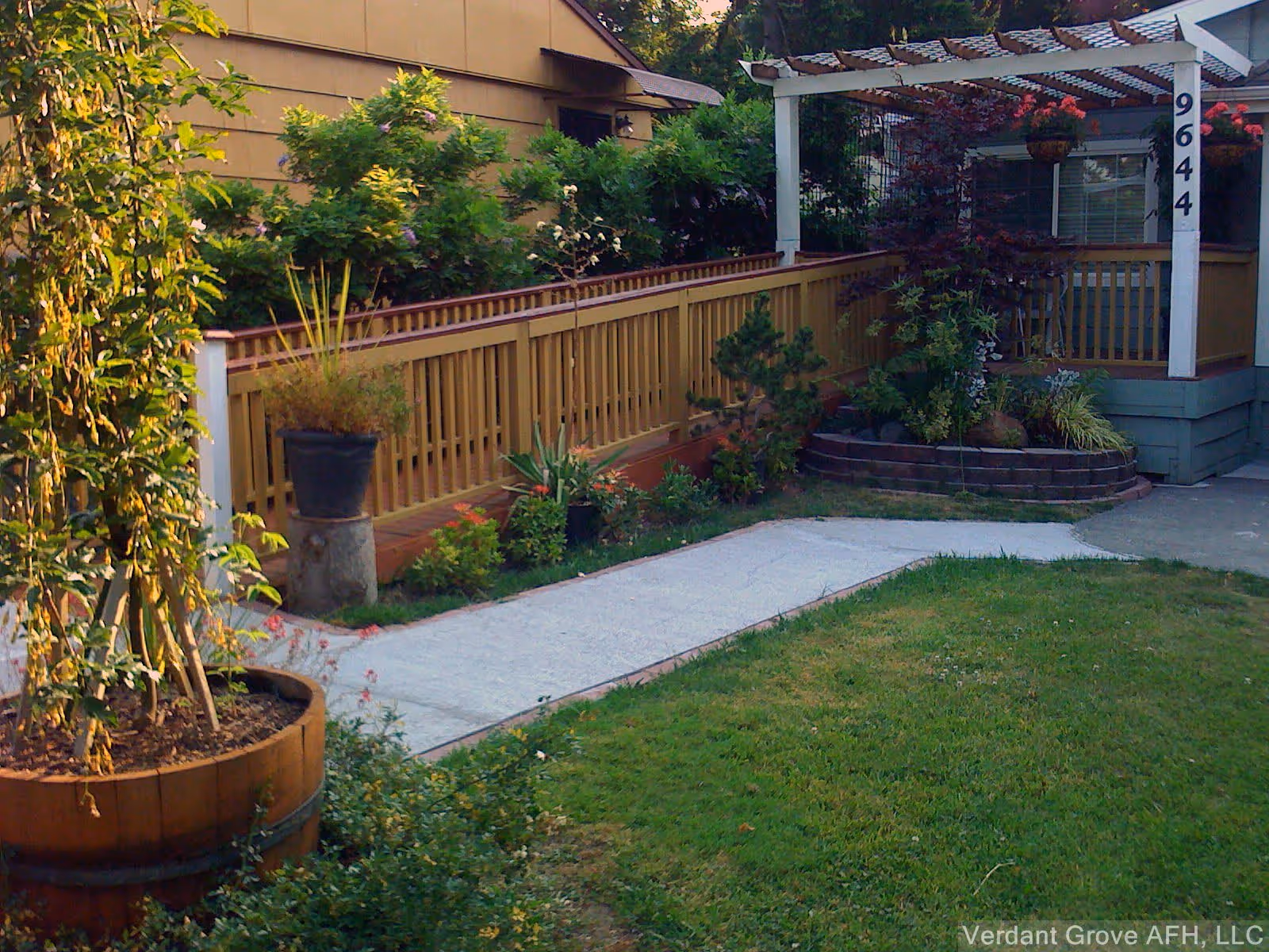 Outdoor view of a garden area with a concrete pathway leading to a porch with a wooden railing and a pergola. The garden has various plants and shrubs, and the porch has the number 9644 displayed on a white post.