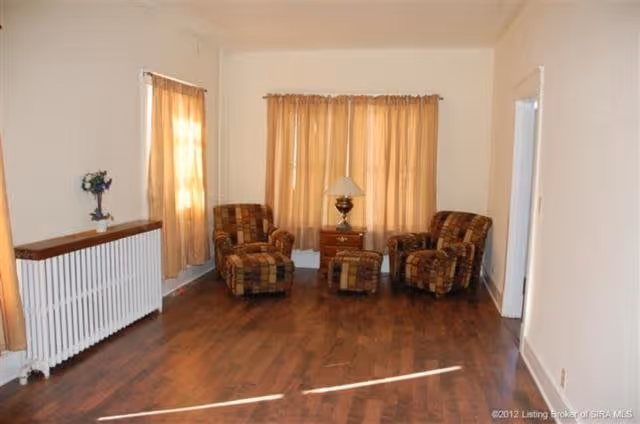 A living room with two patterned armchairs and a matching ottoman arranged around a wooden side table with a lamp. The room has hardwood floors, beige walls, and two windows with beige curtains. There is a white radiator along one wall with a small plant on top.