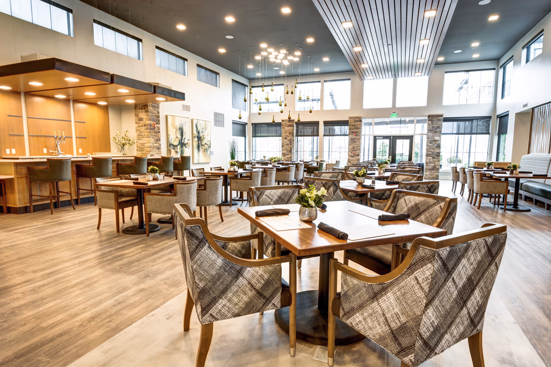 A spacious and well-lit dining room in a senior living facility featuring multiple wooden tables with cushioned chairs arranged neatly. The room has large windows allowing natural light to fill the space, modern pendant lights hanging from the ceiling, and a bar area with high chairs on the left side. The floor is covered with light wood flooring, and small floral centerpieces decorate the tables.
