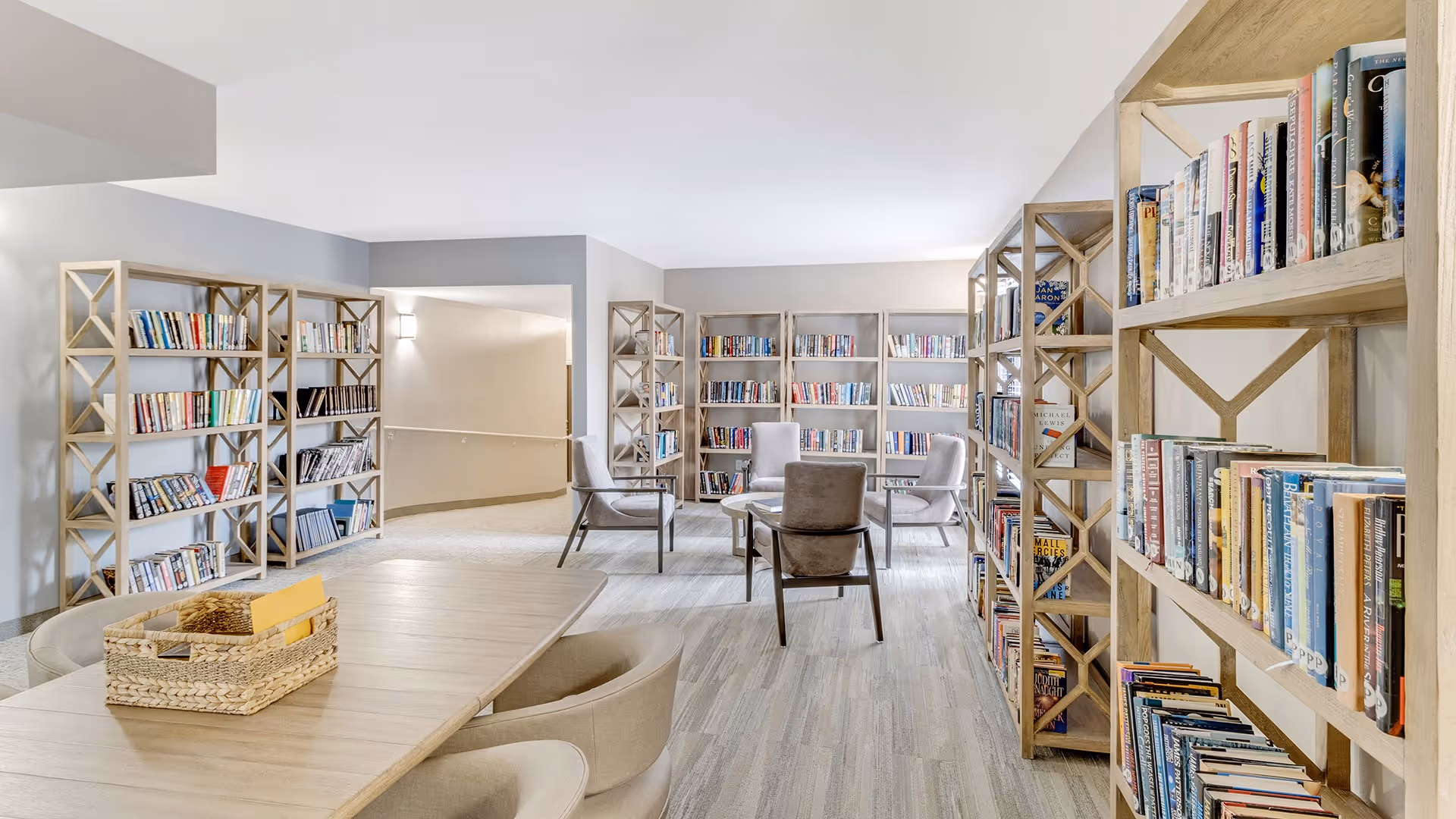 A bright and spacious library room with multiple wooden bookshelves filled with books. There is a large wooden table with beige cushioned chairs around it, and a woven basket on the table. Four additional chairs are arranged in a circle near the back of the room, creating a cozy reading or discussion area. The room has light-colored walls and flooring, with soft lighting.