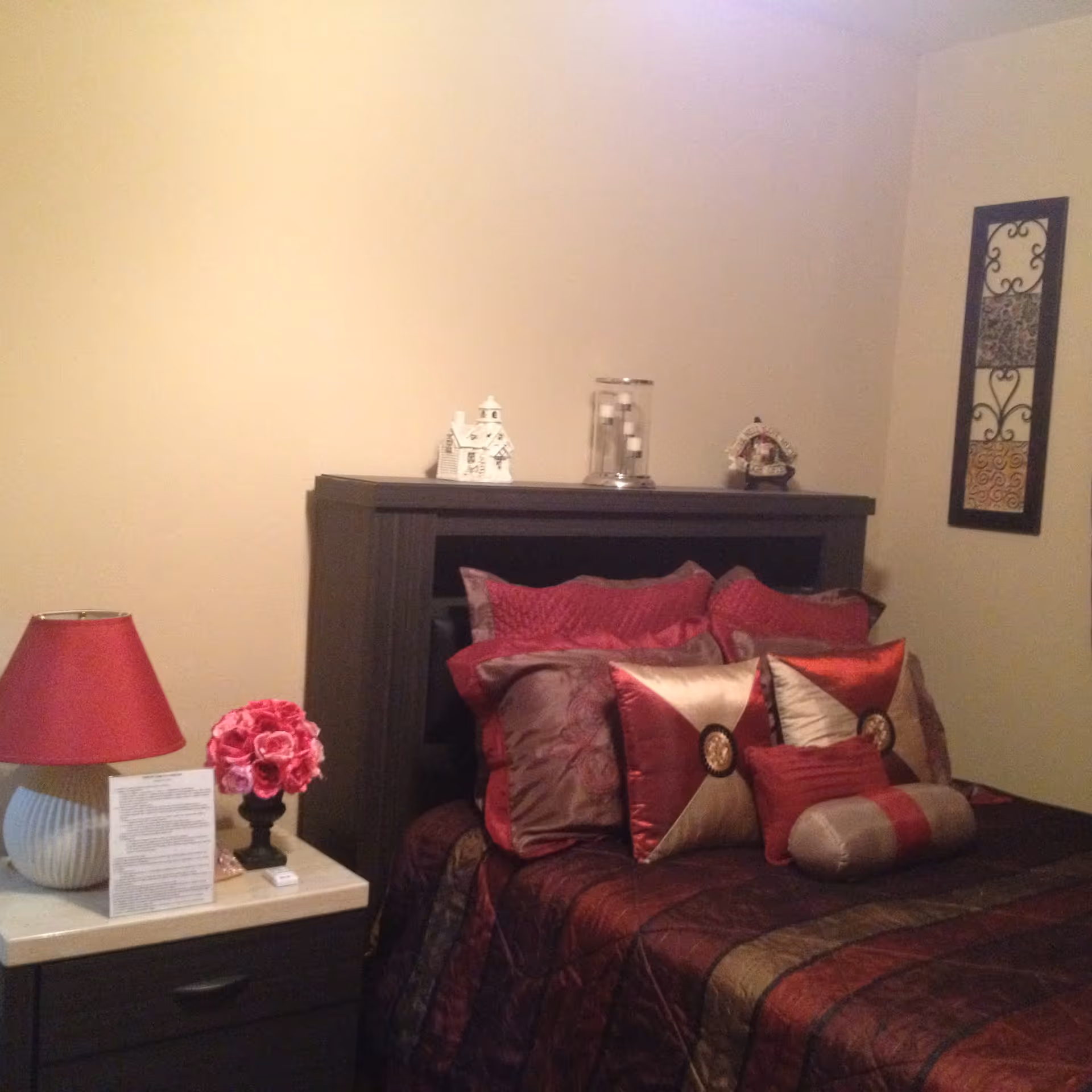 A bedroom with a dark wooden headboard and bed frame. The bed is made with a dark red and brown comforter and several decorative pillows in shades of red, gold, and brown. On the left side, there is a nightstand with a red lampshade, a small bouquet of pink flowers in a black vase, and a piece of paper. On the wall to the right of the bed, there is a decorative wall hanging with intricate patterns.