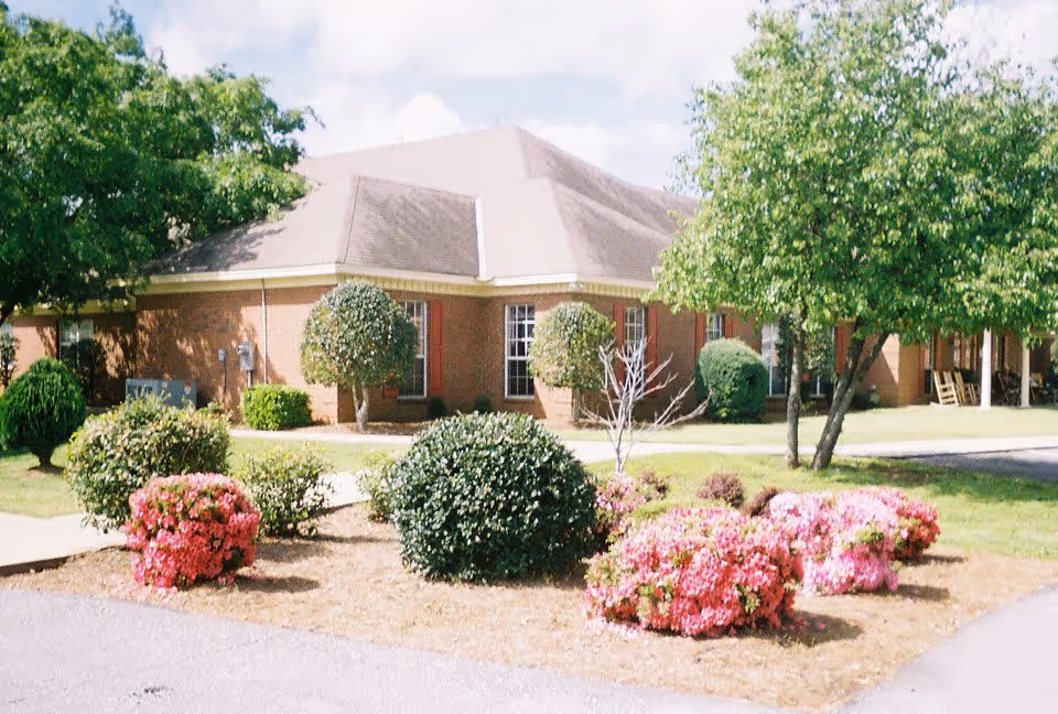 Exterior view of a single-story brick building with a sloped roof, surrounded by well-maintained landscaping including green bushes, pink flowering shrubs, and trees under a partly cloudy sky.