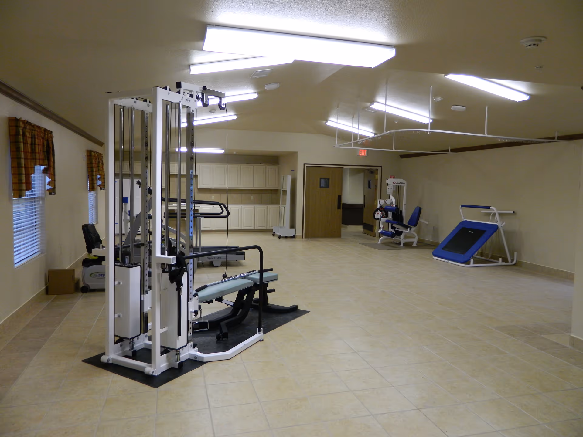 A spacious fitness room in a senior living facility with various exercise equipment including a multi-station gym machine, a recumbent bike, a leg press machine, and a small trampoline. The room has tiled floors, beige walls, fluorescent ceiling lights, and windows with plaid curtains.