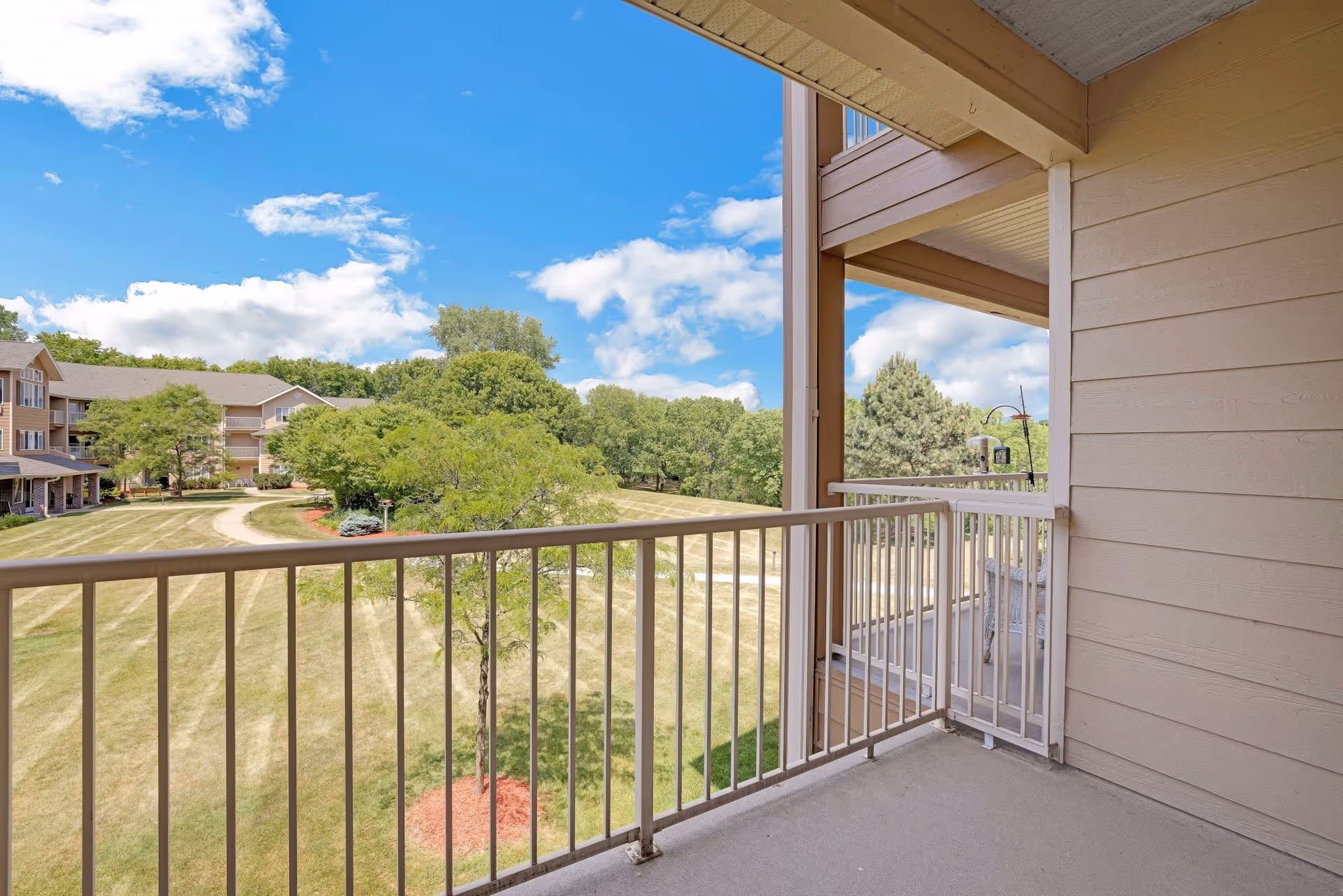 View from a balcony overlooking a grassy area with trees and a pathway, with a multi-story residential building in the background under a partly cloudy blue sky.