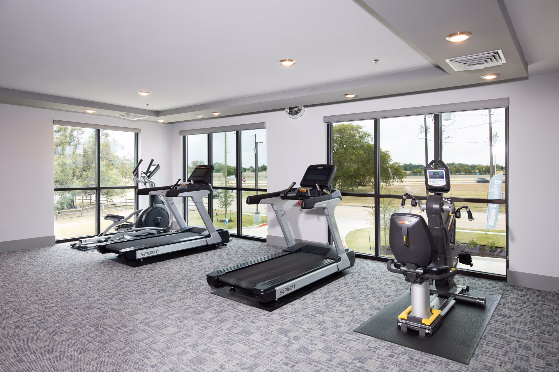 A bright fitness room with treadmills and cardio equipment arranged in front of large windows overlooking the grounds.