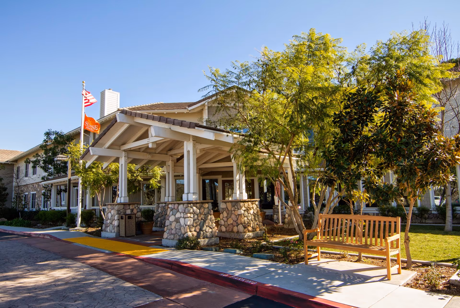 Exterior view of a senior living facility entrance with a covered porch supported by stone pillars, surrounded by trees and greenery. There is a wooden bench on the sidewalk and two flags flying on flagpoles, one of which is the American flag. The sky is clear and blue.