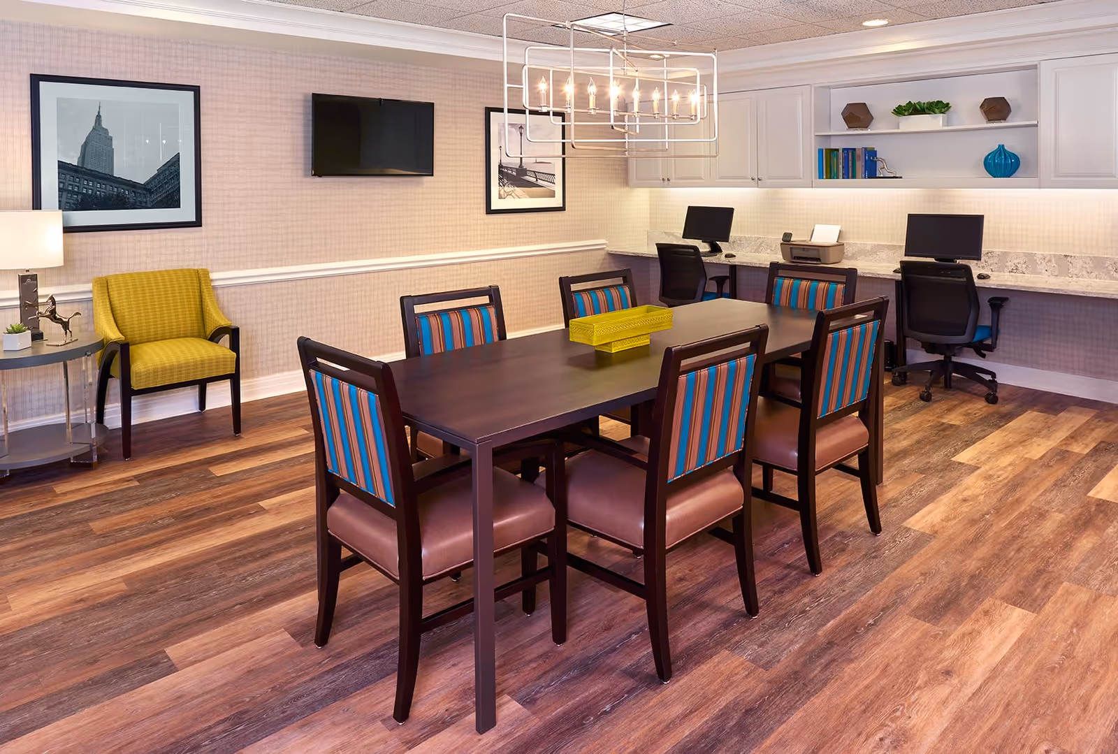 A well-lit room with a dark wooden table surrounded by six chairs with striped blue and brown upholstery. In the background, there is a long counter with two computer workstations and office chairs. The walls are decorated with framed black and white photographs, and a flat-screen TV is mounted on the wall. A modern rectangular chandelier hangs above the table, and a yellow armchair sits next to a small round side table with a lamp and decorative items. The floor has a wood-like finish.