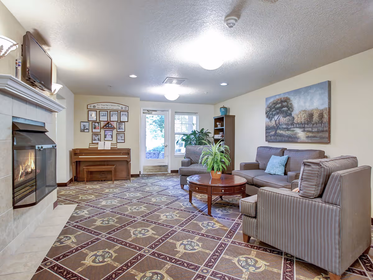 A cozy communal living room with sofas, armchairs, a round coffee table, a fireplace, and a piano under framed artwork.