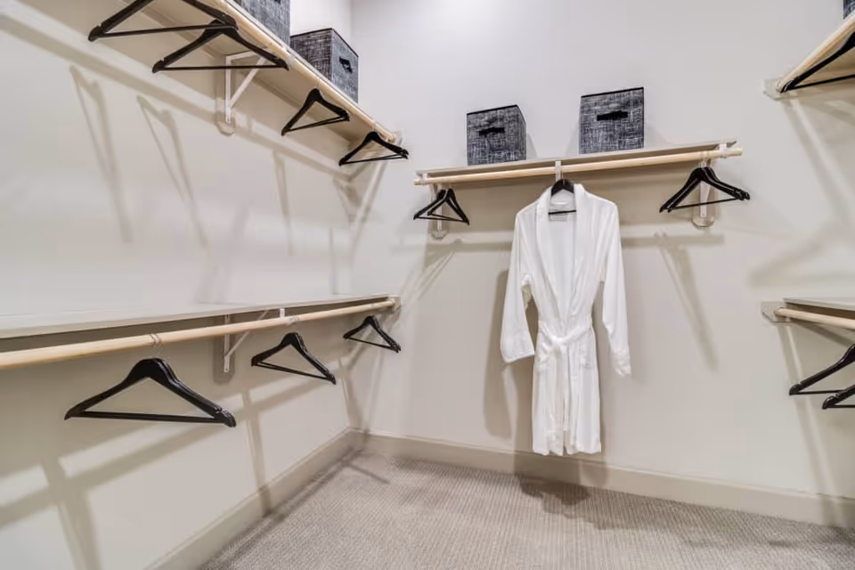 A spacious walk-in closet with multiple wooden hanging rods and black hangers. Two gray storage bins are placed on the upper shelves, and a white bathrobe hangs on one of the rods. The closet has light-colored walls and carpeted flooring.