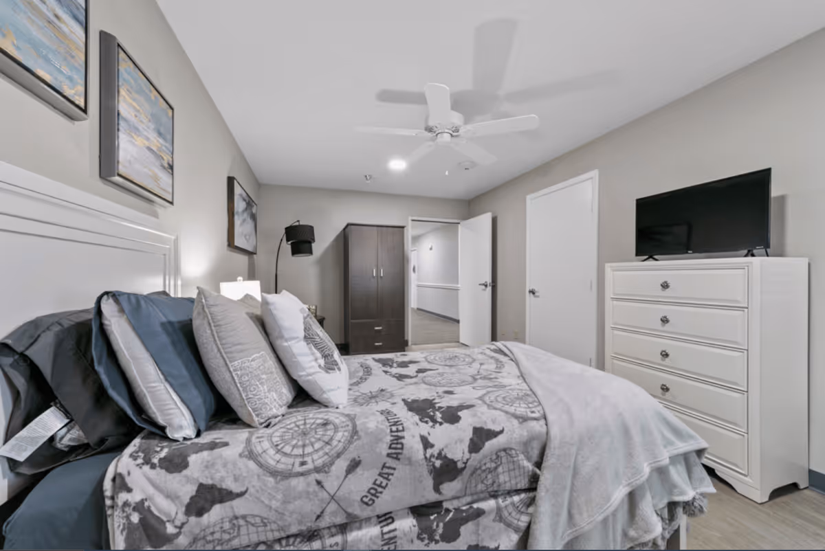 A neatly made bed with multiple pillows and a blanket featuring a world map design in a bedroom. The room has light-colored walls, a ceiling fan, a white dresser with a TV on top, a dark wardrobe, and two doors, one of which is open showing a hallway.
