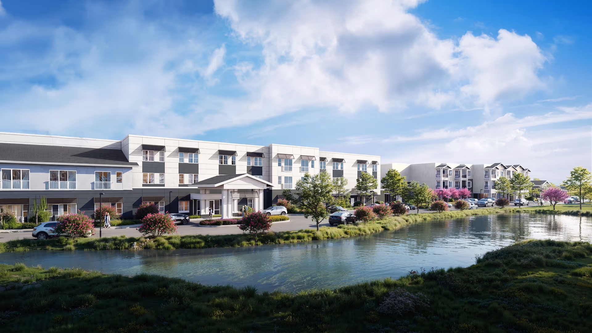 Exterior front view of a modern three-story senior living building beside a pond with parked cars and landscaped grounds under a blue sky.