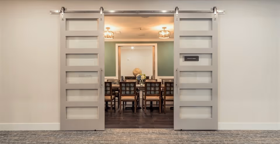 View through partially open sliding barn doors into a private dining room with a long table and multiple chairs. The room has a green accent wall and two ceiling light fixtures.