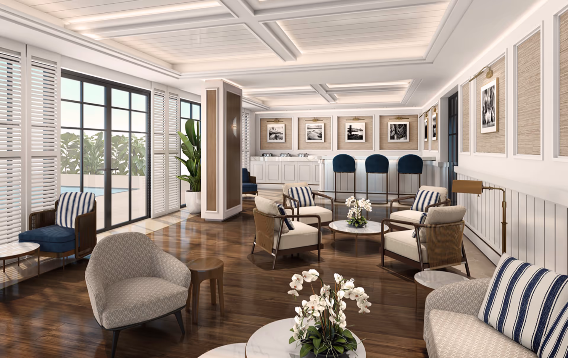 A spacious and elegant living room area with wooden flooring, multiple comfortable chairs with cushions, small round tables, and a bar counter with three blue bar stools. The room has large windows with white shutters allowing natural light and a view of greenery outside. The walls are decorated with framed black and white photographs and soft lighting fixtures.