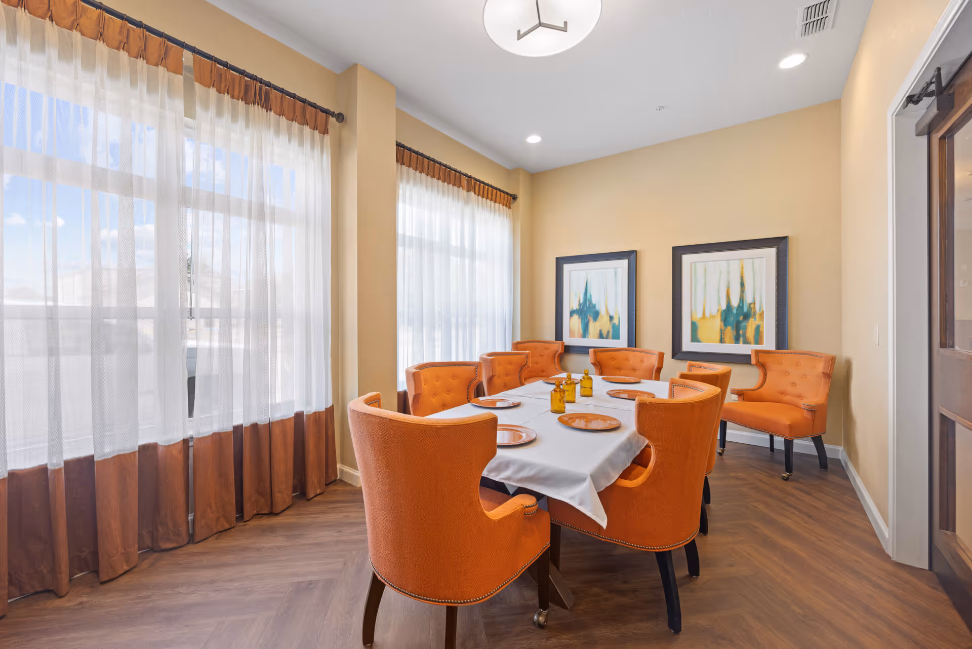 A dining room with a rectangular table covered with a white tablecloth, surrounded by eight orange upholstered chairs. The table is set with orange plates and three small amber glass bottles. The room has large windows with sheer white and brown curtains, light beige walls, two abstract paintings in black frames, and wood flooring.