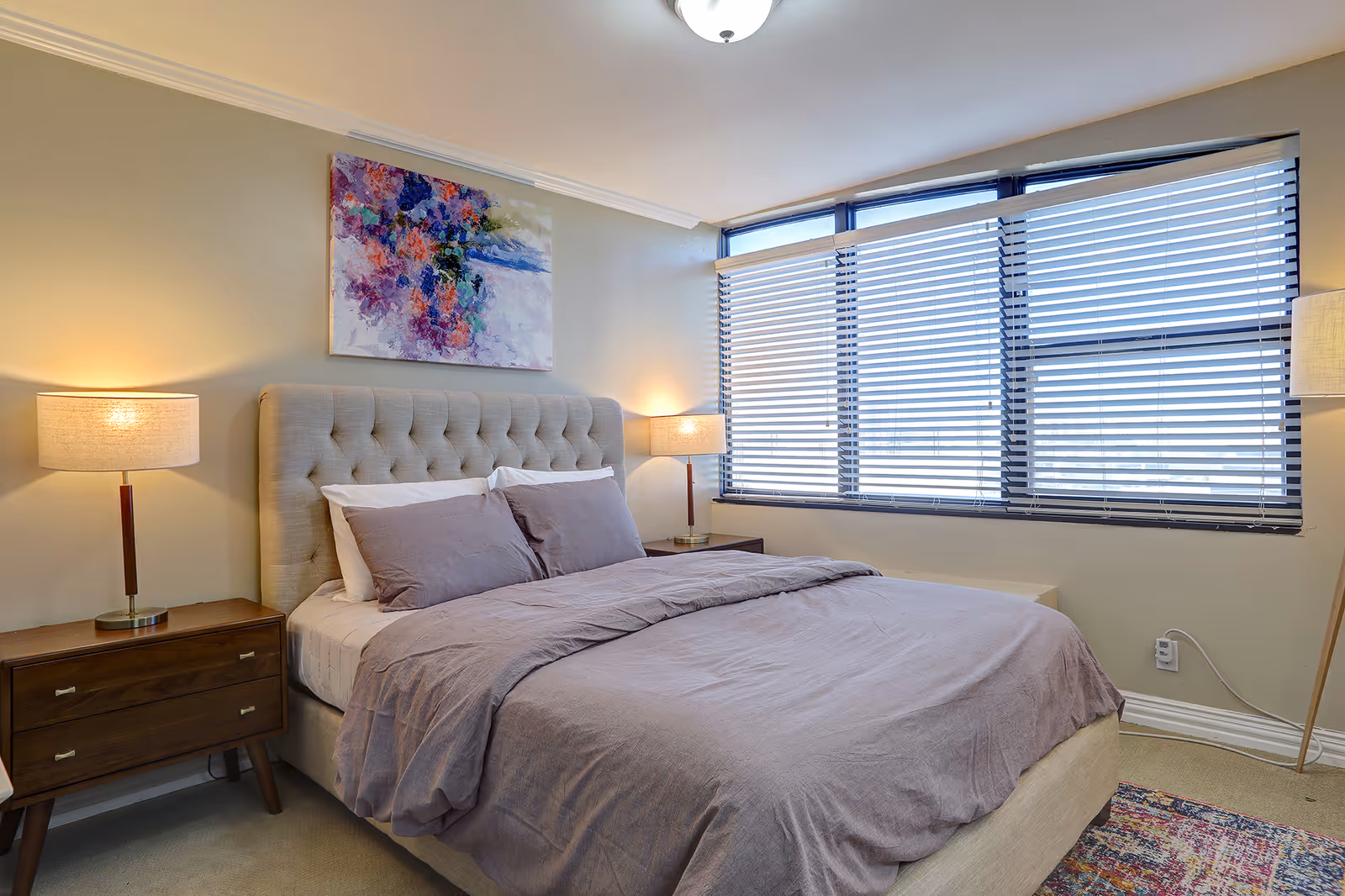 A cozy bedroom with a large bed featuring a tufted headboard, gray bedding, and two pillows. There are two wooden nightstands on either side of the bed, each with a lit lamp. A colorful abstract painting hangs above the bed. The room has a large window with horizontal blinds partially closed, allowing natural light to enter. A patterned rug is partially visible on the floor.