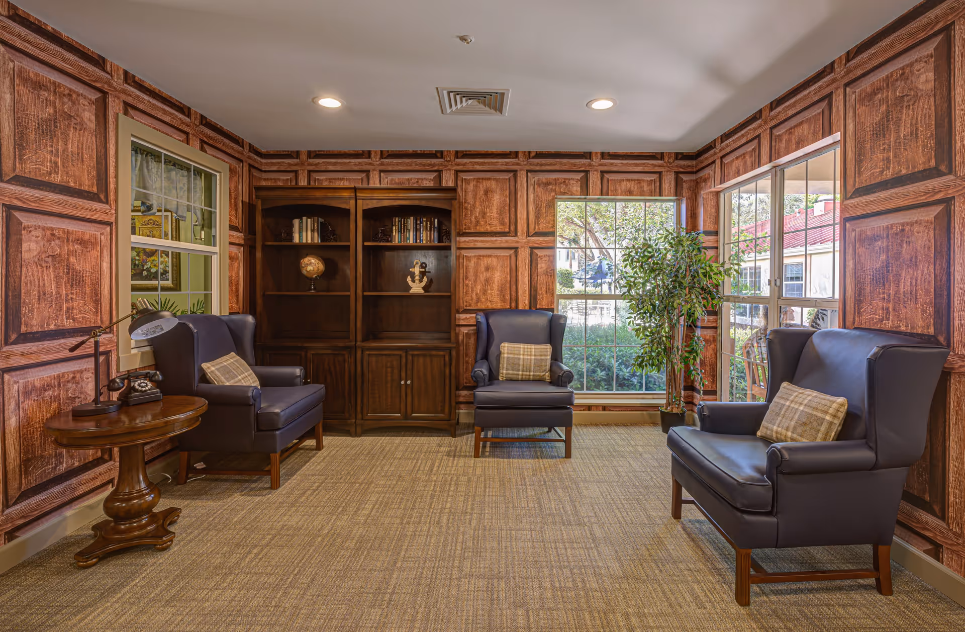 A cozy sitting area with three dark leather armchairs, each with a plaid cushion, arranged around a wooden side table with a vintage telephone and lamp. The room features wood-paneled walls, a carpeted floor, a wooden bookshelf with decorative items, and large windows letting in natural light with a view of greenery outside.