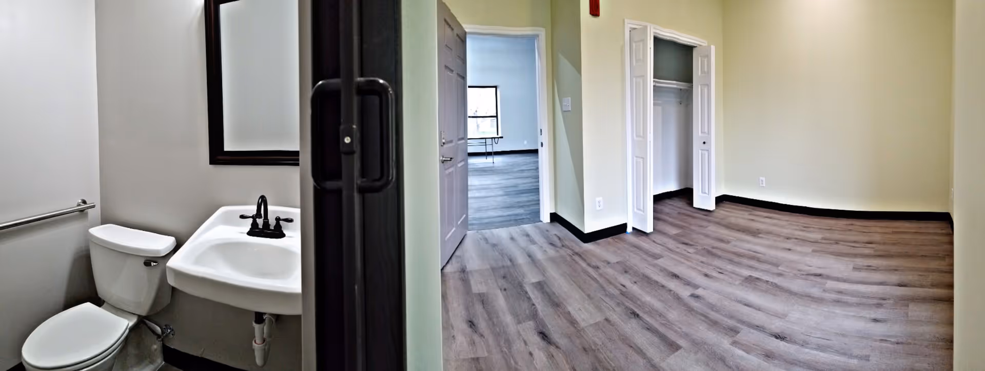 View of a small bathroom with a toilet, sink, and mirror on the left side, and an empty room with wood flooring, a closet with open bi-fold doors, and a doorway leading to another room with a window and table in the background on the right side.