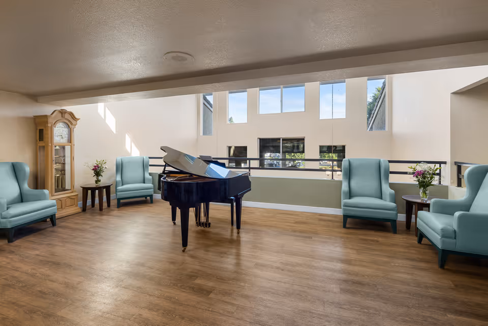 A spacious senior living common area with a black grand piano in the center, surrounded by four light blue armchairs. There are two small round tables with flower arrangements next to the chairs. A tall wooden grandfather clock is positioned against the left wall. Large windows on the far wall allow natural light to fill the room, highlighting the wood flooring.