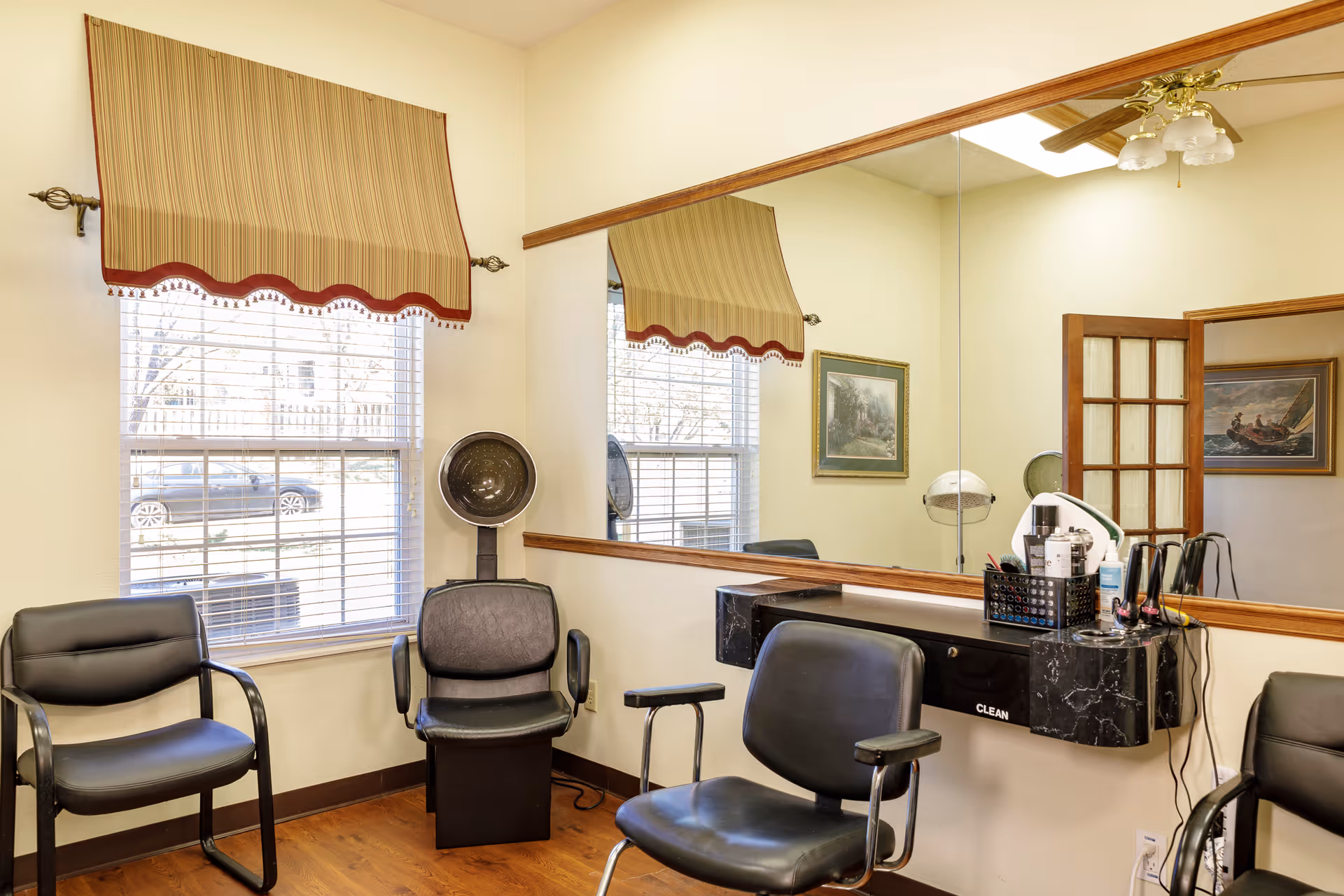 Interior of a small salon or hair styling area with three black chairs, a large mirror on the wall, a window with striped valance, and hair drying equipment. The room has light-colored walls and wooden flooring.