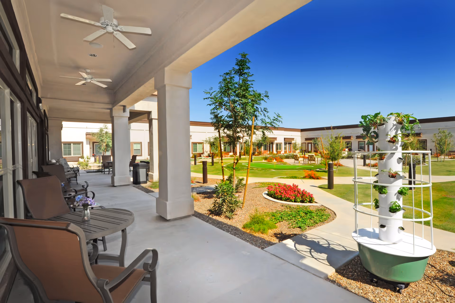 Outdoor patio area at Sky Ridge Senior Living with several chairs and tables under a covered walkway. The patio overlooks a landscaped garden with green grass, small trees, flower beds, and a vertical garden planter. The sky is clear and blue.