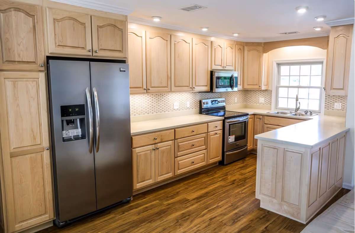 Bright kitchen with stainless steel refrigerator and stove, light wood cabinets, and a peninsula countertop near a window.