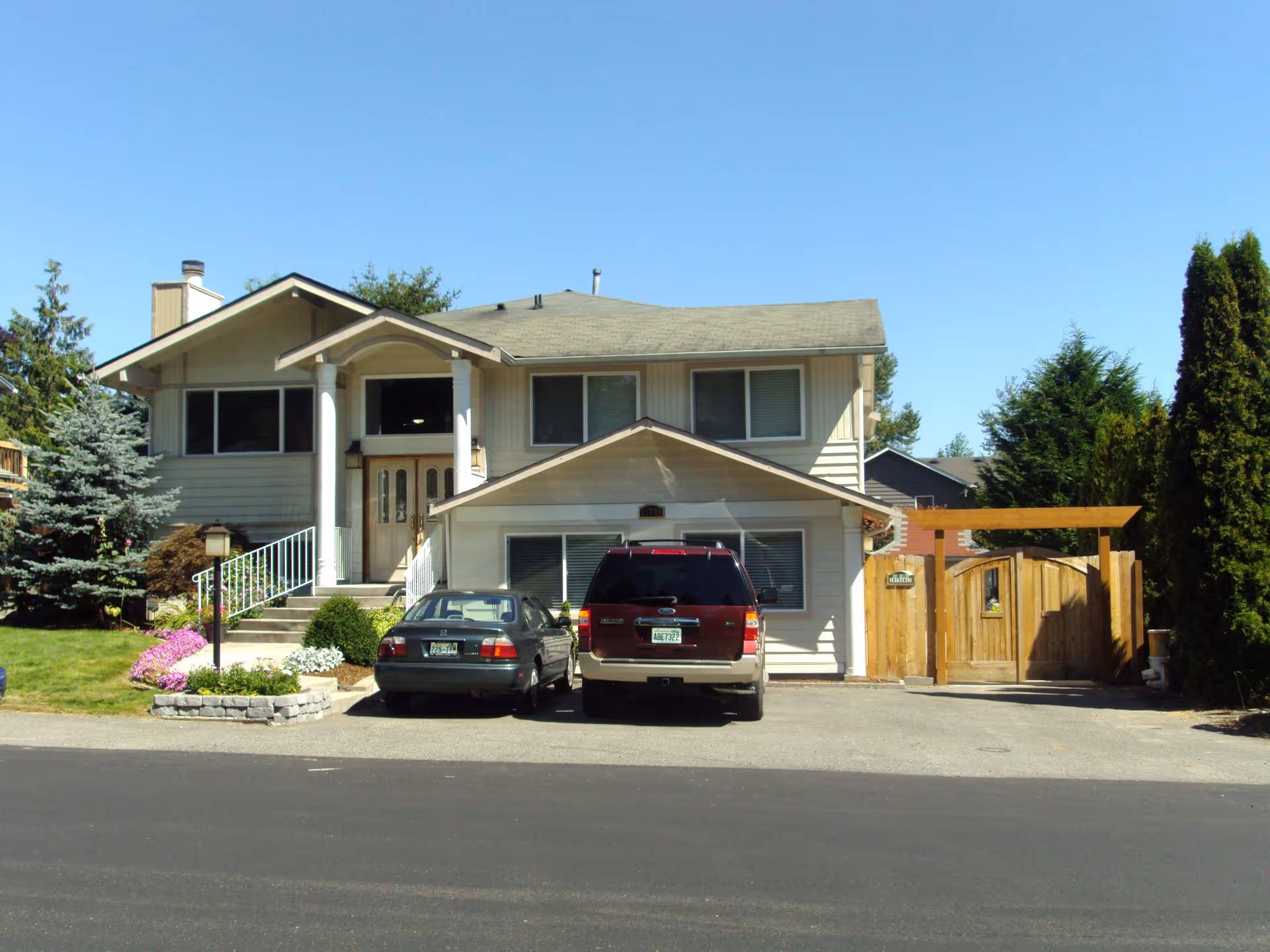 Front exterior view of a two-story residential building with a driveway containing two parked cars. The house has a light-colored facade, a small staircase leading to the front door, and a wooden gate on the right side. There are trees and shrubs around the property under a clear blue sky.