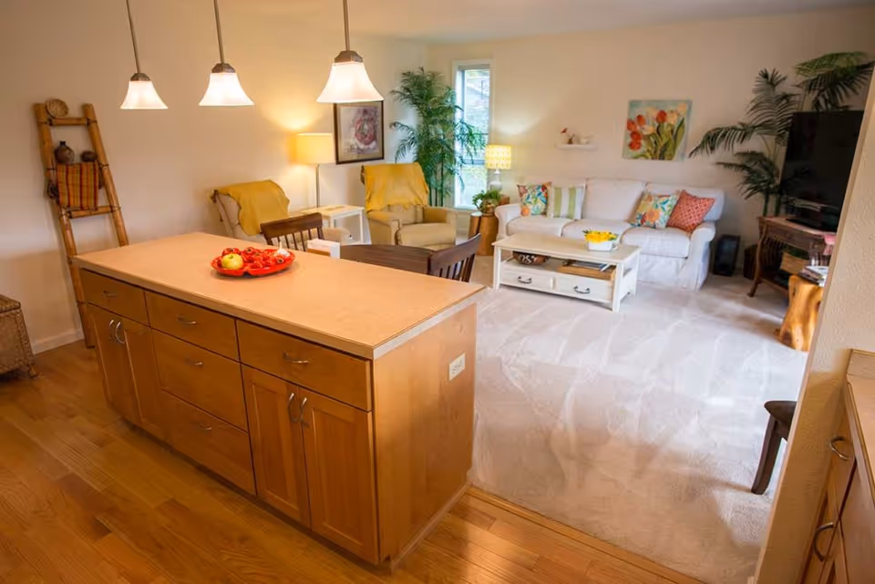 A bright and cozy living room and kitchen area in a senior living facility. The kitchen features a wooden island with drawers and cabinets, topped with a plate of fruit. The living room has a white sofa with colorful pillows, two beige armchairs with yellow throws, a white coffee table, a large TV on a wooden stand, and decorative plants and artwork on the walls. The floor is a combination of hardwood and carpet.