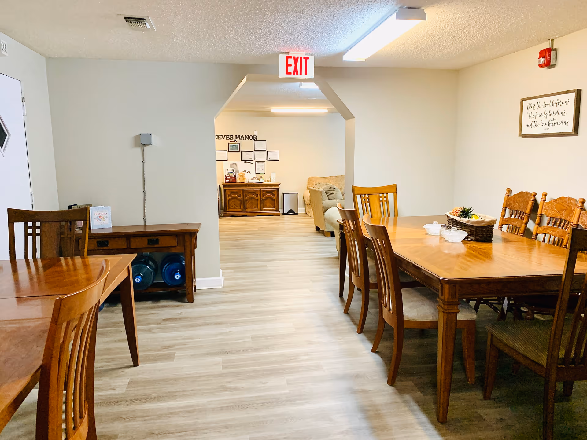 Bright communal dining area with wooden tables and chairs, an exit sign, and a seating area visible through an archway.