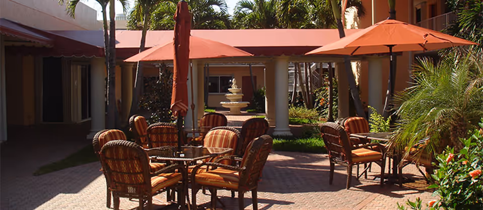 Outdoor courtyard with wicker tables and chairs under orange umbrellas, palm trees, and a central fountain in front of a colonnade.