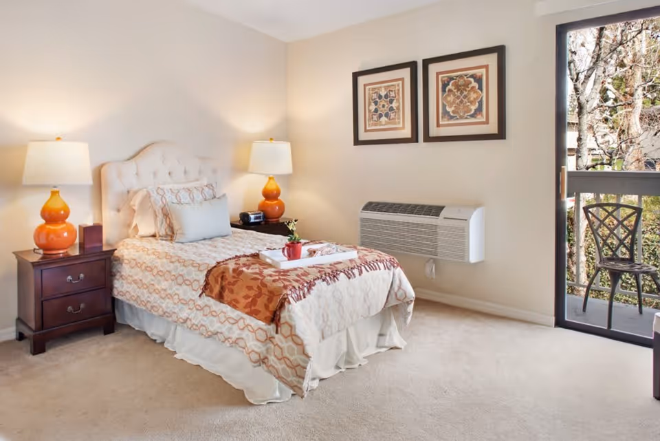 A cozy bedroom with a single bed featuring patterned bedding and a decorative throw. There are two bedside tables, each with an orange lamp. Above the bed are two framed artworks. A wall-mounted air conditioning unit is visible, and a sliding glass door opens to a small balcony with a metal chair and a view of trees outside.