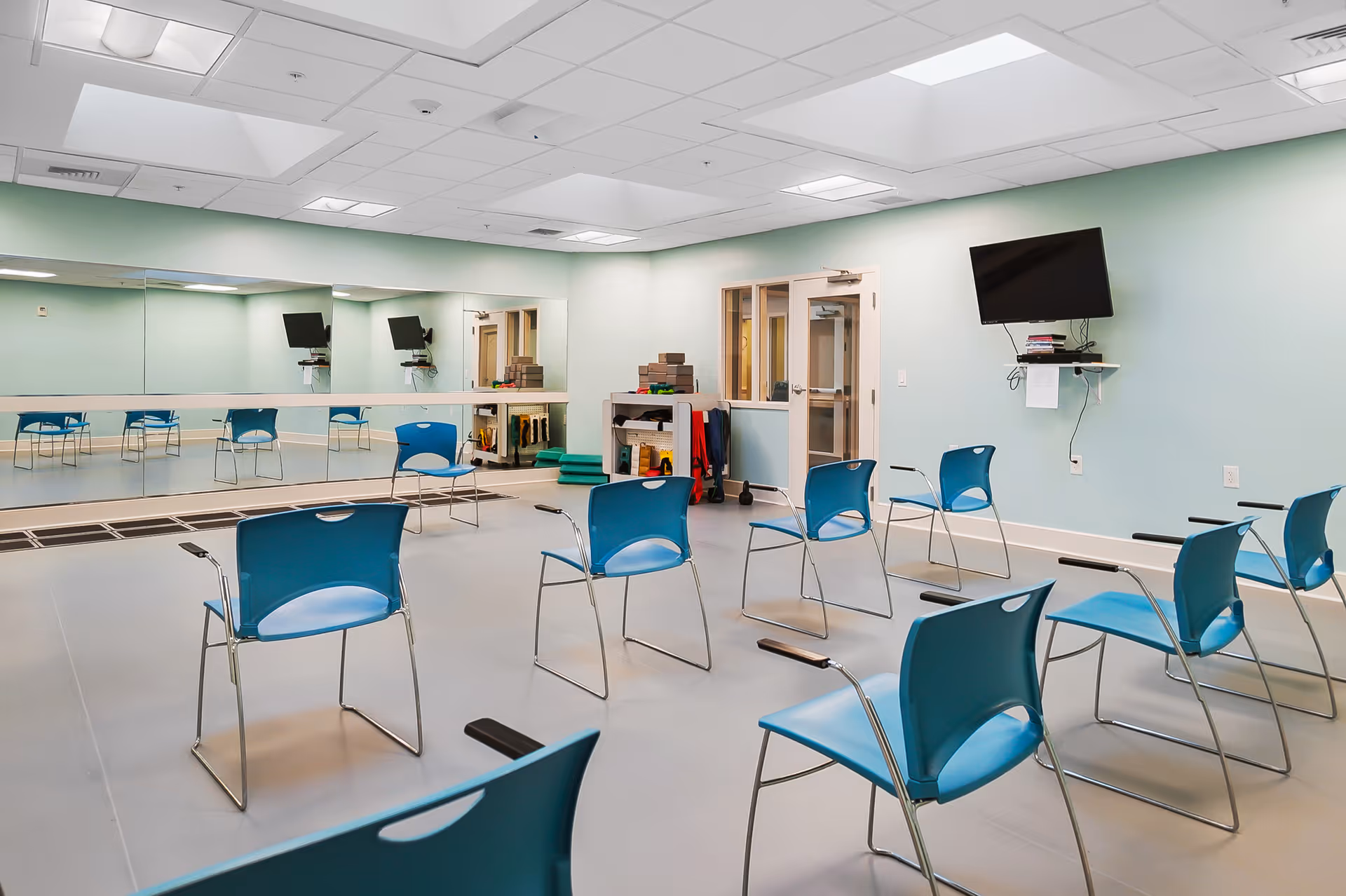 Spacious exercise/activity room with blue chairs arranged in rows facing mirrors and a wall-mounted TV.