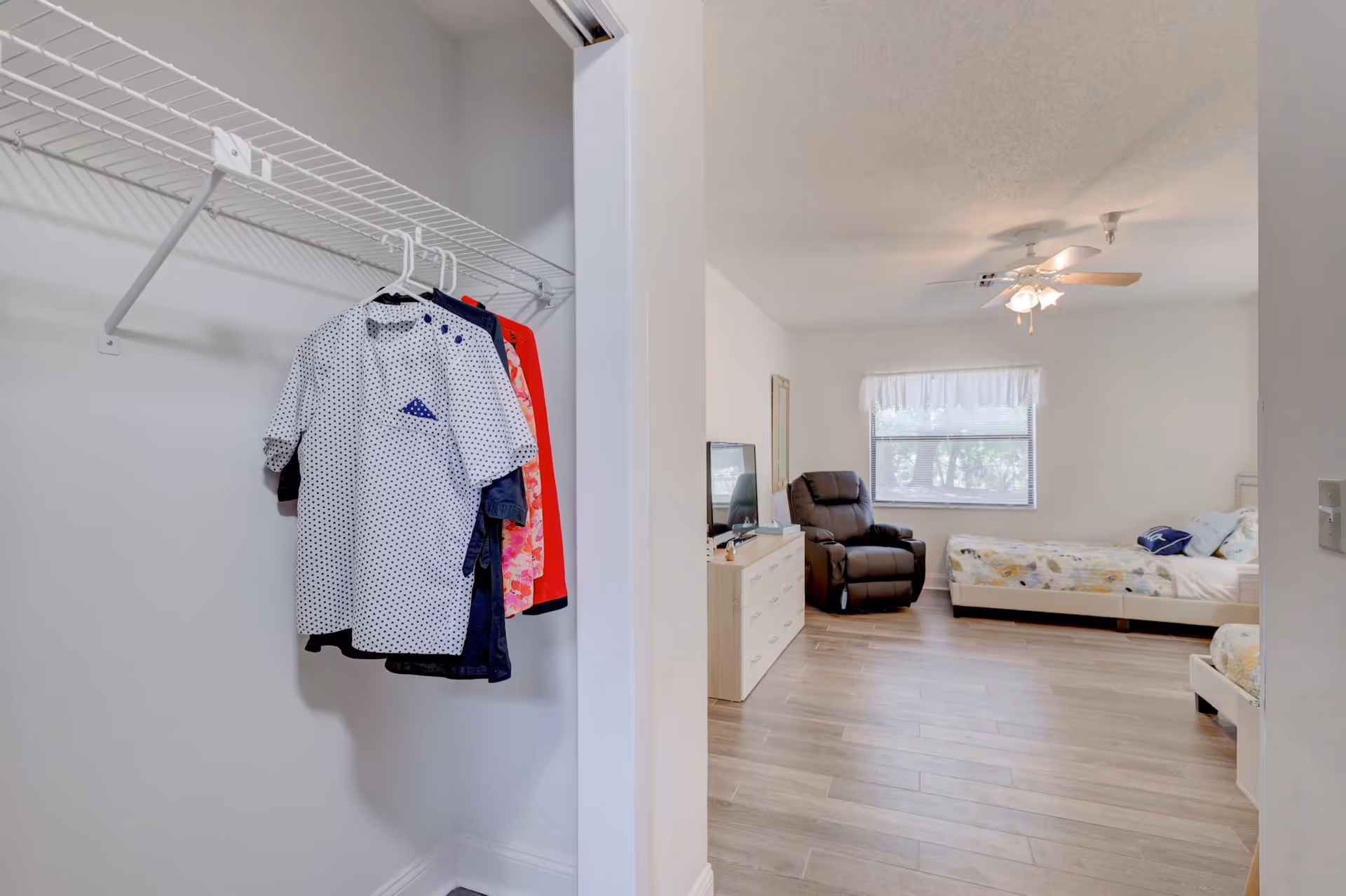 View from a closet into a bright furnished bedroom with beds, a recliner, dresser and ceiling fan.