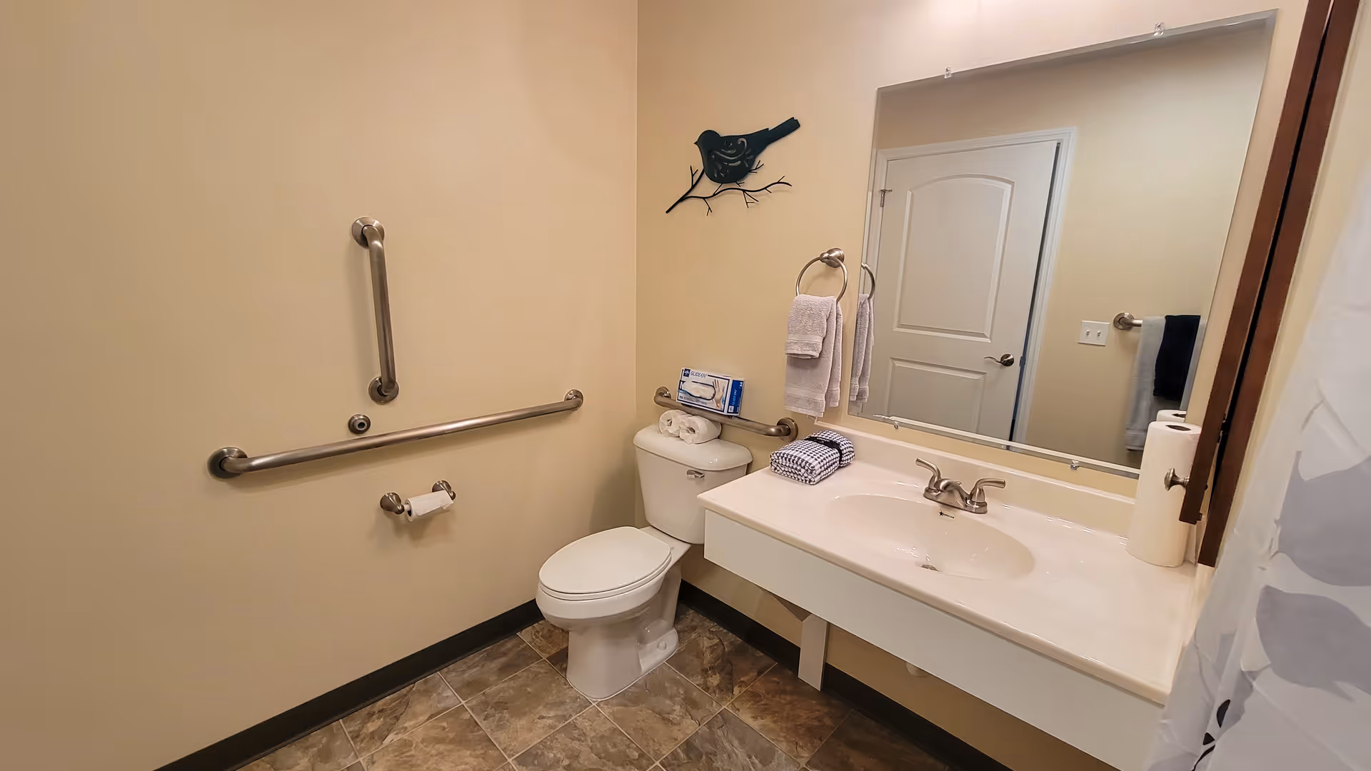 A clean and accessible bathroom with beige walls and tiled floor. The bathroom features a white toilet with grab bars on the adjacent walls, a white sink with a countertop, a large mirror above the sink, and a towel ring holding two white towels. A decorative black bird wall art is mounted above the toilet. A roll of paper towels is on the sink counter, and a shower curtain with a leaf pattern is partially visible on the right side.