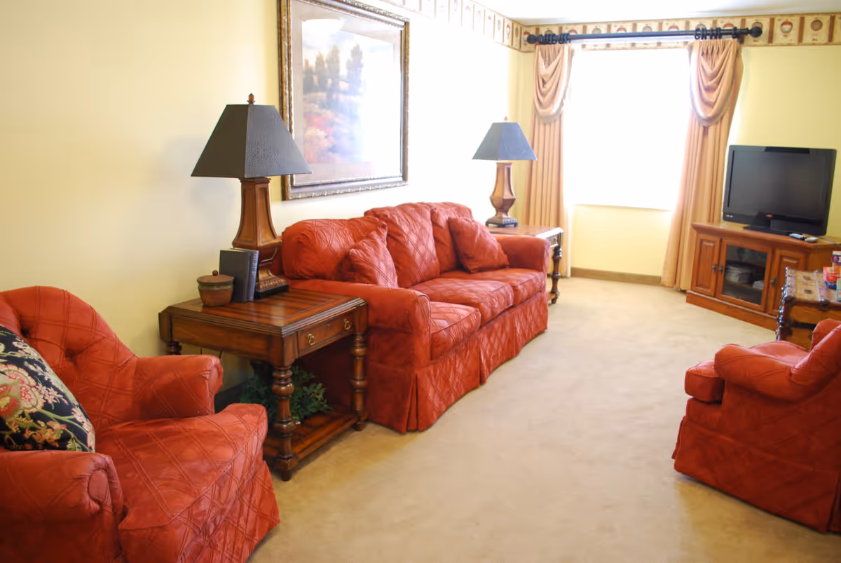 Bright living room with red upholstered sofa and armchairs, wooden side tables with lamps, and a TV near a curtained window.